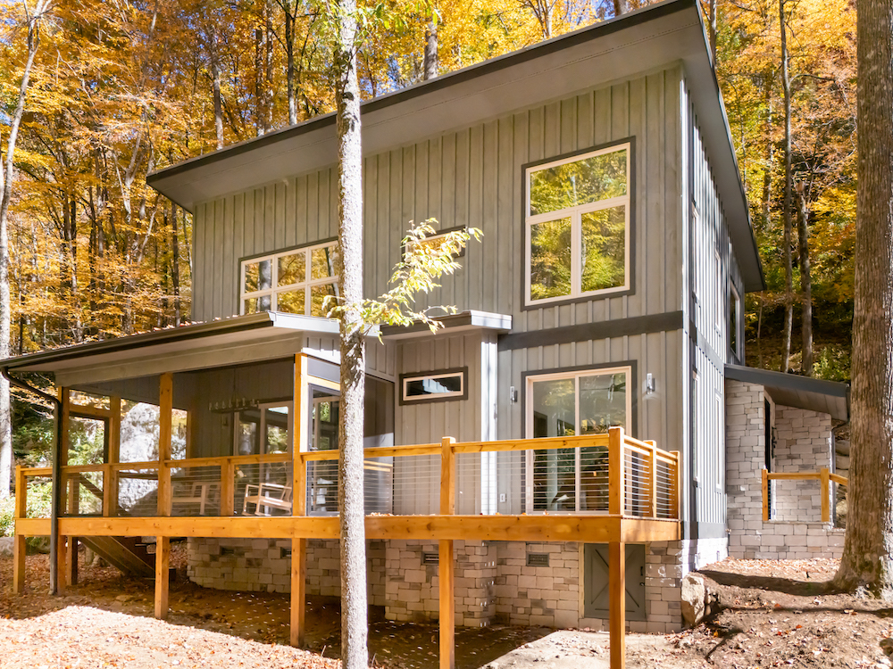 A recently completed custom mountain home by Powell Construction, set into hardwoods at peak fall color on the Highlands-Cashiers Plateau, with board-and-batten siding, stone-clad foundation, and a covered porch with cable railing and Adirondack chairs