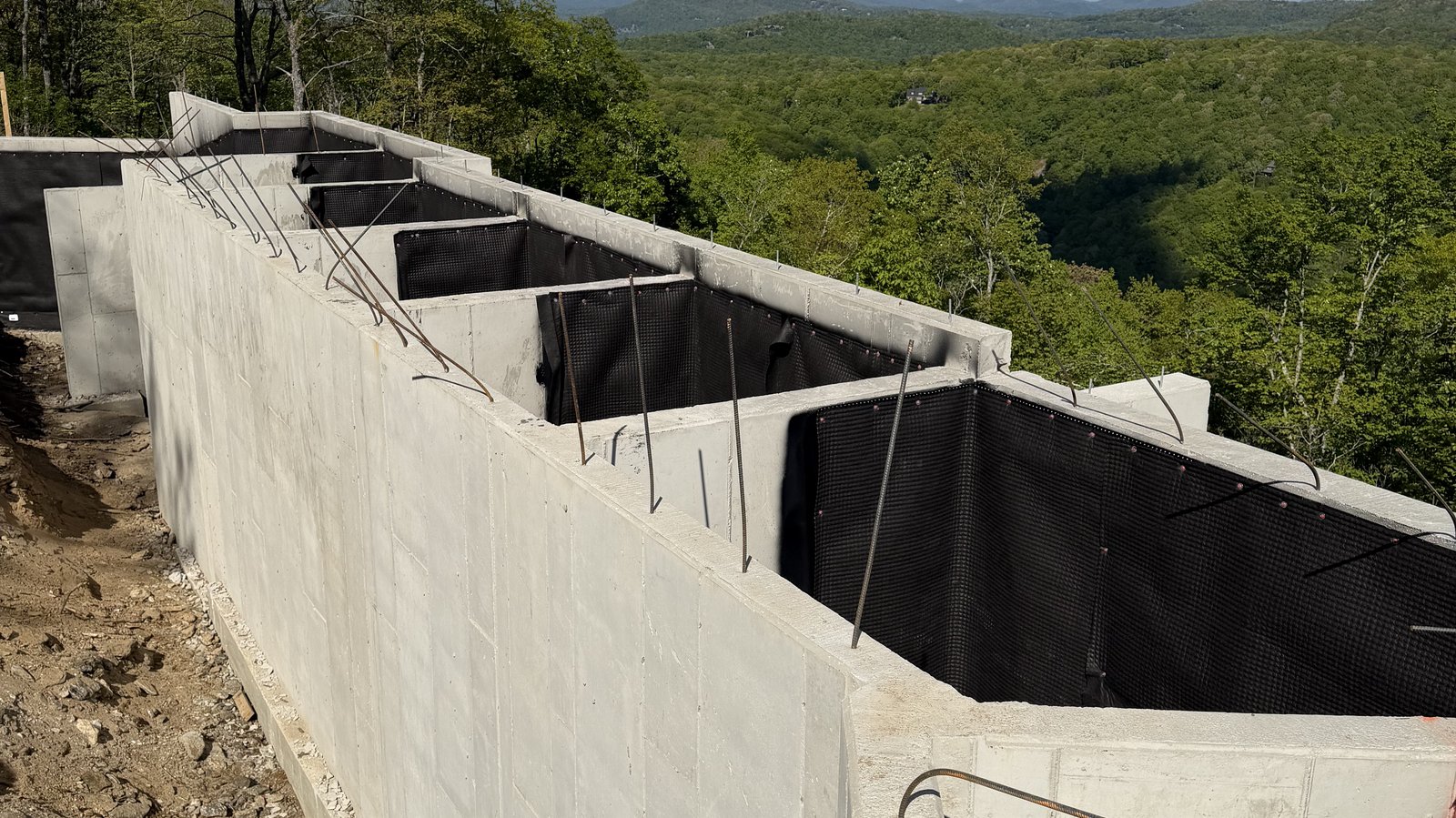 A poured concrete foundation mid-build on a Powell Construction custom home in the Highlands-Cashiers Plateau, with exposed rebar, waterproofing membrane, and the Blue Ridge ridgeline visible in the distance